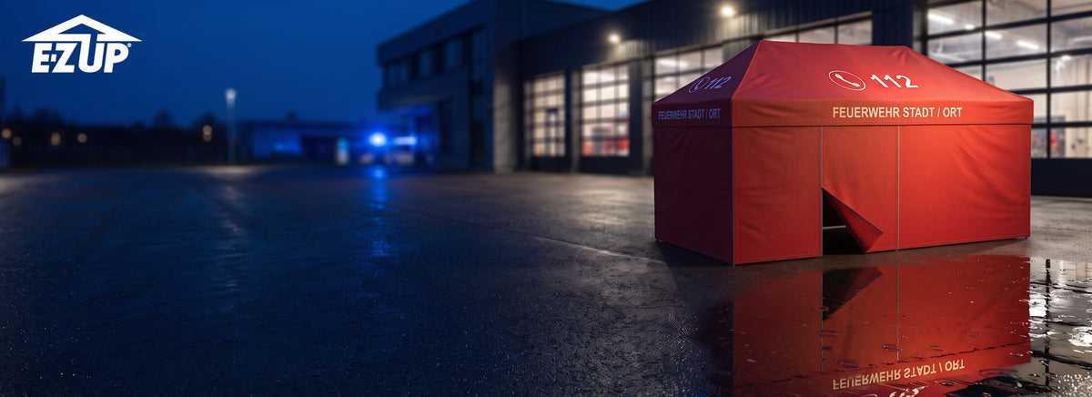 Roter E-Z UP Feuerwehr-Pavillon 3x6 m mit Dachdruck „112“ und „FEUERWEHR STADT / ORT“ vor einem Feuerwehrhaus am Abend